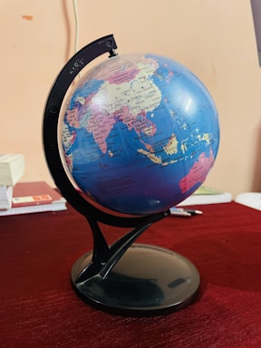 A globe displaying several countries including India, China, and surrounding regions is mounted on a black stand. It is positioned on a table with a dark red tablecloth. Some books and a pen are partially visible in the background, adding an academic or educational ambience.