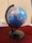 A globe displaying several countries including India, China, and surrounding regions is mounted on a black stand. It is positioned on a table with a dark red tablecloth. Some books and a pen are partially visible in the background, adding an academic or educational ambience.