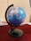 A globe displaying several countries including India, China, and surrounding regions is mounted on a black stand. It is positioned on a table with a dark red tablecloth. Some books and a pen are partially visible in the background, adding an academic or educational ambience.