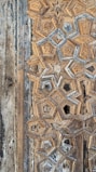 A close-up of intricate geometric wood carvings forming star-like patterns. The aged wood displays a combination of beige, brown, and hints of gray, giving it an antique appearance. The texture and detail emphasize craftsmanship and historical aesthetic.