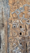 A close-up of intricate geometric wood carvings forming star-like patterns. The aged wood displays a combination of beige, brown, and hints of gray, giving it an antique appearance. The texture and detail emphasize craftsmanship and historical aesthetic.