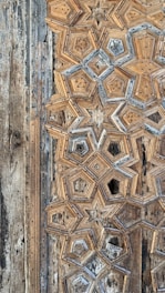 A close-up of intricate geometric wood carvings forming star-like patterns. The aged wood displays a combination of beige, brown, and hints of gray, giving it an antique appearance. The texture and detail emphasize craftsmanship and historical aesthetic.