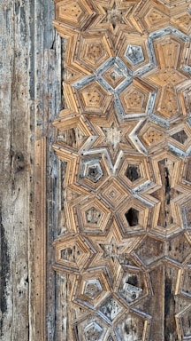 A close-up of intricate geometric wood carvings forming star-like patterns. The aged wood displays a combination of beige, brown, and hints of gray, giving it an antique appearance. The texture and detail emphasize craftsmanship and historical aesthetic.
