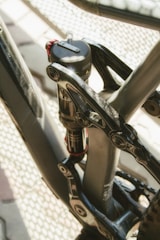 Close-up of a bicycle's rear suspension system, highlighting the metallic components and the shock absorber. The image captures intricate details of the mechanism, including bolts and the mounting structure, with sunlight reflecting off the surfaces.