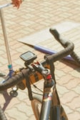 A close-up view of a bicycle handlebar equipped with a digital device, possibly a GPS or speedometer. There is a strap for holding items attached to the handlebar. In the background, part of a scooter and a person's hand are visible on a paved surface with a ramp or platform.