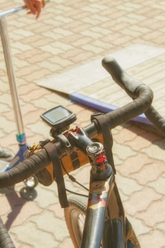 A close-up view of a bicycle handlebar equipped with a digital device, possibly a GPS or speedometer. There is a strap for holding items attached to the handlebar. In the background, part of a scooter and a person's hand are visible on a paved surface with a ramp or platform.