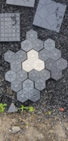 A collection of hexagonal and square paving tiles laid out on a surface with small black stones in the background. The center features a light-colored hexagonal tile surrounded by darker tiles, creating a geometric pattern. Sparse greenery surrounds the bottom edge.
