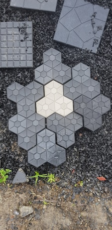 A collection of hexagonal and square paving tiles laid out on a surface with small black stones in the background. The center features a light-colored hexagonal tile surrounded by darker tiles, creating a geometric pattern. Sparse greenery surrounds the bottom edge.