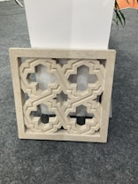 A decorative stone panel featuring an intricate geometric pattern is positioned against a white rectangular structure. The panel displays symmetrical cut-out designs and is placed on a gray carpeted floor.