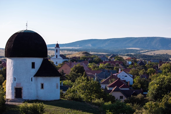 A historic village scene features a round white church with a dark, dome-shaped roof in the foreground. The background includes rolling hills under a clear blue sky. A mix of colorful rooftops and lush greenery can be seen throughout the village.