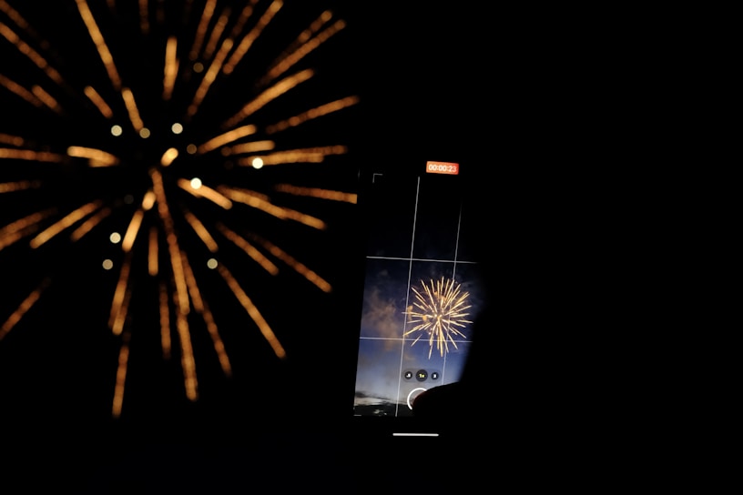 A large firework burst is captured through the screen of a smartphone, which is recording the event. The explosion illuminates the dark background with golden and bright hues, creating an impressive display. The smartphone screen also shows a grid overlay for alignment, with a duration timer at the top.