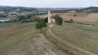 A rural landscape featuring a small stone church with a tall bell tower situated on a gentle hill. The surrounding area includes expansive fields and rolling hills with scattered trees. A few farm buildings can be seen in the distance, and the sky is overcast.