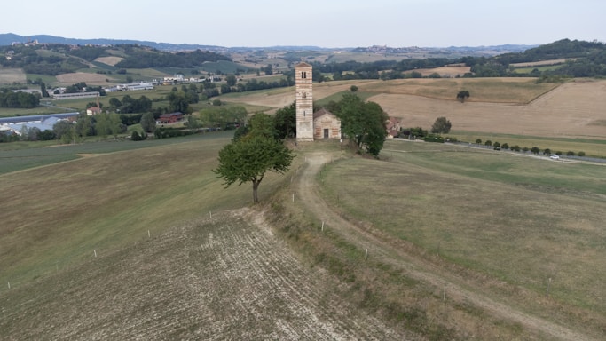 A rural landscape featuring a small stone church with a tall bell tower situated on a gentle hill. The surrounding area includes expansive fields and rolling hills with scattered trees. A few farm buildings can be seen in the distance, and the sky is overcast.
