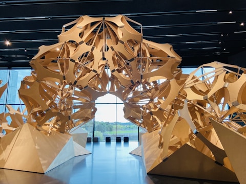 A geometric wooden structure with interlocking shapes placed in an indoor space with large glass windows in the background. The structure forms an intricate design with triangular and curved elements, casting shadows on the floor.