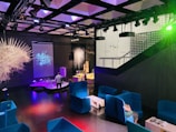 A modern indoor space features vibrant blue seating areas with armchairs and small tables. Several colorful installations and illuminated displays create a futuristic atmosphere. The room is dimly lit with prominent lighting effects, and a projection of an abstract design is visible on one wall. A person is interacting with one of the exhibits.