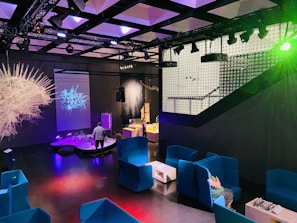 A modern indoor space features vibrant blue seating areas with armchairs and small tables. Several colorful installations and illuminated displays create a futuristic atmosphere. The room is dimly lit with prominent lighting effects, and a projection of an abstract design is visible on one wall. A person is interacting with one of the exhibits.