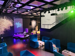 A modern indoor space features vibrant blue seating areas with armchairs and small tables. Several colorful installations and illuminated displays create a futuristic atmosphere. The room is dimly lit with prominent lighting effects, and a projection of an abstract design is visible on one wall. A person is interacting with one of the exhibits.