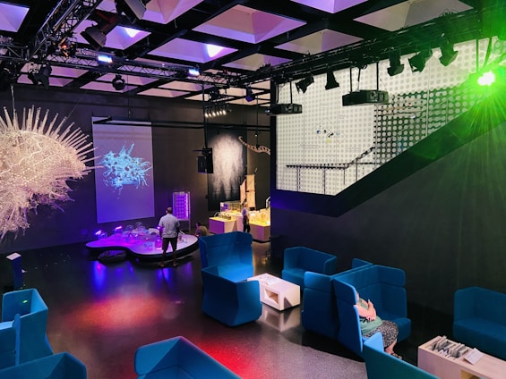 A modern indoor space features vibrant blue seating areas with armchairs and small tables. Several colorful installations and illuminated displays create a futuristic atmosphere. The room is dimly lit with prominent lighting effects, and a projection of an abstract design is visible on one wall. A person is interacting with one of the exhibits.