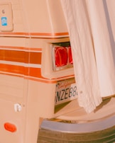 A close-up view of the back of a vintage vehicle, partially covered by a curtain. The image reveals a taillight, a California license plate, and decorative striped lines in shades of orange and beige. The overall aesthetic is retro, with a warm, nostalgic vibe.