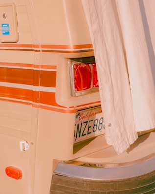 A close-up view of the back of a vintage vehicle, partially covered by a curtain. The image reveals a taillight, a California license plate, and decorative striped lines in shades of orange and beige. The overall aesthetic is retro, with a warm, nostalgic vibe.