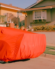 A car covered with a bright red cover is parked on the side of a street in front of a house. The house is green with a white-framed window and various potted plants and garden decorations lining the front yard. Neighboring houses and part of the street are also visible.