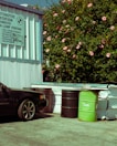 An automotive station with a corrugated metal wall featuring a sign for various car services. A black car is partially visible, parked near several large barrels, one of which is green with 'SAM' written on it. In the background, a lush bush is adorned with numerous pink flowers.