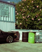 An automotive station with a corrugated metal wall featuring a sign for various car services. A black car is partially visible, parked near several large barrels, one of which is green with 'SAM' written on it. In the background, a lush bush is adorned with numerous pink flowers.