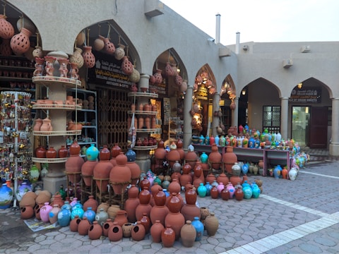 A marketplace stall displaying an array of colorful and traditional pottery items, including vases, jugs, and lamps, arranged neatly on the ground and shelves. The pottery features intricate designs and varies in color from terracotta to bright blues and pinks, under an arcade structure with arches.