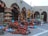 A marketplace stall displaying an array of colorful and traditional pottery items, including vases, jugs, and lamps, arranged neatly on the ground and shelves. The pottery features intricate designs and varies in color from terracotta to bright blues and pinks, under an arcade structure with arches.