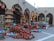 A marketplace stall displaying an array of colorful and traditional pottery items, including vases, jugs, and lamps, arranged neatly on the ground and shelves. The pottery features intricate designs and varies in color from terracotta to bright blues and pinks, under an arcade structure with arches.