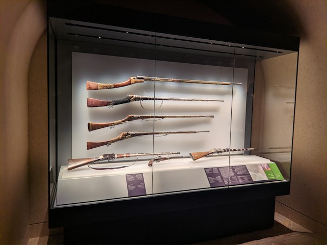 A museum display case containing five antique rifles, each aligned horizontally on a white background. The rifles vary in size and design, with intricate details visible on their wooden and metal parts. Plaques with descriptions are positioned below the rifles.