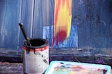 A close-up view of a paint can filled with black paint and a brush inside. Next to the can is a multicolored palette. The background features a partially painted surface with broad strokes of yellow and red over a blue canvas.
