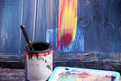 A close-up view of a paint can filled with black paint and a brush inside. Next to the can is a multicolored palette. The background features a partially painted surface with broad strokes of yellow and red over a blue canvas.