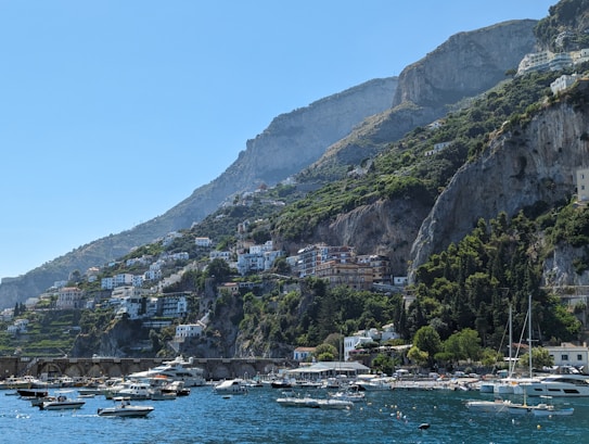 The image features a picturesque coastal landscape with rugged cliffs and lush greenery. A cluster of white buildings with terracotta roofs is nestled into the hillside. In the foreground, a calm, deep blue sea is dotted with small boats and yachts. The sky is clear and blue, adding to the tranquil atmosphere.