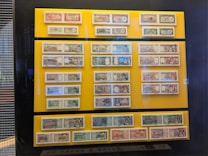 A collection of various banknotes is displayed behind glass on a bright yellow background. The banknotes are arranged in rows and columns and feature different colors, designs, and portraits. The display is mounted on a wall and surrounded by a dark frame.