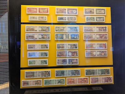 A collection of various banknotes is displayed behind glass on a bright yellow background. The banknotes are arranged in rows and columns and feature different colors, designs, and portraits. The display is mounted on a wall and surrounded by a dark frame.