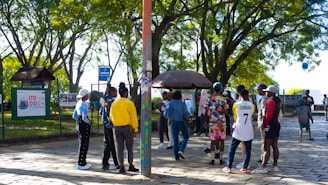 A group of people gathers in a park under leafy trees. They are interacting near a kiosk or stand surrounded by greenery and paved ground. Some individuals are wearing colorful clothing, and one person is wearing a number 7 sports jersey. There's a mixture of standing and walking activities happening in the scene.