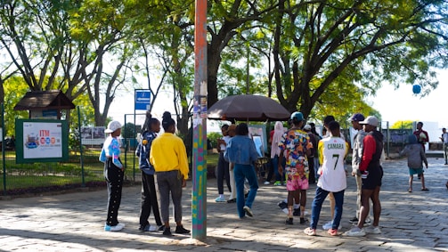 A group of people gathers in a park under leafy trees. They are interacting near a kiosk or stand surrounded by greenery and paved ground. Some individuals are wearing colorful clothing, and one person is wearing a number 7 sports jersey. There's a mixture of standing and walking activities happening in the scene.