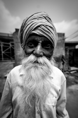 An elderly man with a long white beard and a turban on his head, standing in an outdoor setting with a blurred background of buildings and sky. His face appears weathered and expressive, suggesting wisdom and experience.