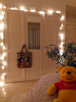 A cozy room features string lights around white shutter doors, a decorative bag with Mickey Mouse design hanging from the door, a potted plant, and a plush toy bear wearing a red shirt. The room has a warm and inviting atmosphere.