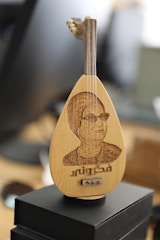 An intricately crafted miniature stringed instrument featuring a carved image of a person on its body with some text beneath the carving. The instrument is placed on a dark box, and the background is blurred giving a depth of field effect.