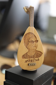 An intricately crafted miniature stringed instrument featuring a carved image of a person on its body with some text beneath the carving. The instrument is placed on a dark box, and the background is blurred giving a depth of field effect.