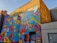 A vibrant mural on the side of a building with geometric patterns and shapes in various bright colors, featuring the text 'CREATIVITY IS IN ALL OF US'. The mural is located at the London Graphic Centre. The background includes a yellow brick wall and other urban elements such as doors and a lamp.