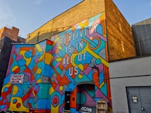 A vibrant mural on the side of a building with geometric patterns and shapes in various bright colors, featuring the text 'CREATIVITY IS IN ALL OF US'. The mural is located at the London Graphic Centre. The background includes a yellow brick wall and other urban elements such as doors and a lamp.