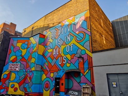 A vibrant mural on the side of a building with geometric patterns and shapes in various bright colors, featuring the text 'CREATIVITY IS IN ALL OF US'. The mural is located at the London Graphic Centre. The background includes a yellow brick wall and other urban elements such as doors and a lamp.