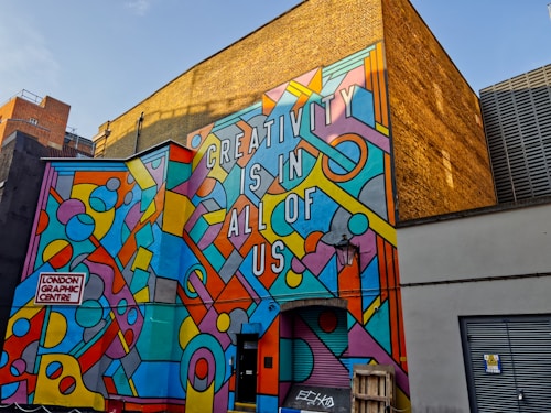 A vibrant mural on the side of a building with geometric patterns and shapes in various bright colors, featuring the text 'CREATIVITY IS IN ALL OF US'. The mural is located at the London Graphic Centre. The background includes a yellow brick wall and other urban elements such as doors and a lamp.