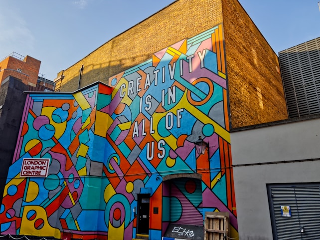 A vibrant mural on the side of a building with geometric patterns and shapes in various bright colors, featuring the text 'CREATIVITY IS IN ALL OF US'. The mural is located at the London Graphic Centre. The background includes a yellow brick wall and other urban elements such as doors and a lamp.