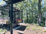 A garden setting featuring a structure with a metal frame and mesh walls, adorned with flower boxes filled with colorful pink and purple blooms. Surrounding the structure is a lush forest with tall trees and ample greenery. The ground near the structure is covered with mulch, featuring a few small plants.
