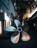 A large metallic ship propeller is positioned outdoors on a paved walkway. The surroundings include a building with windows and a tree casting shadows against the wall. The scene has a warm, golden light filtering through the leaves, creating a calming atmosphere.