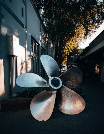 A large metallic ship propeller is positioned outdoors on a paved walkway. The surroundings include a building with windows and a tree casting shadows against the wall. The scene has a warm, golden light filtering through the leaves, creating a calming atmosphere.